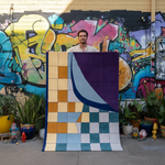 Person holding a colorful rug in front of a graffiti-covered wall.