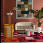 A stylish living room featuring a Vermilion Grid Hand Tufted Area Rug. The contemporary wool rug, with its vibrant red, orange, and pink geometric pattern, covers the floor, complementing the modern decor. A sleek sofa and elegant table sit atop the large area rug, perfect for enhancing any living room space.