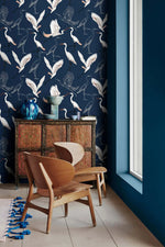 Navy wallpaper with white cranes in corner with carved wood cabinet, blue vases and wood chair