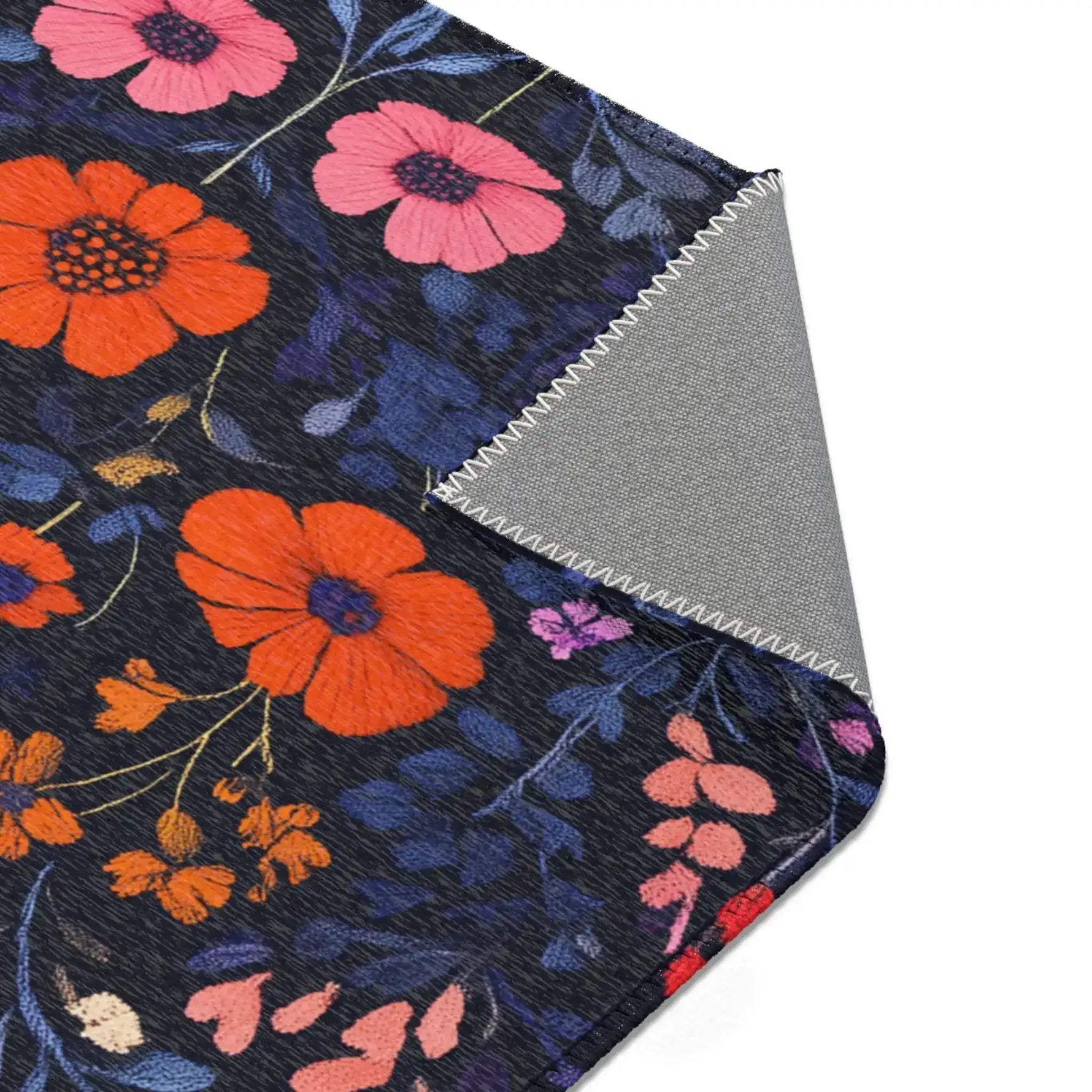 Corner detail of Vibrant Floral Embroidered Accent Rug folded flat, large orange cosmos and pink wildflowers on navy ground, grey backing visible