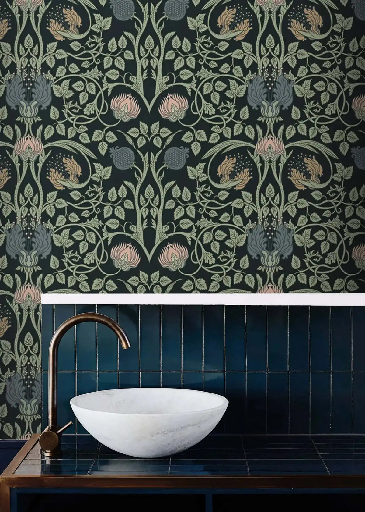 Vine and flower wallpaper in bathroom above dark blue vertical tiles with white marble basin and bronze gooseneck faucet