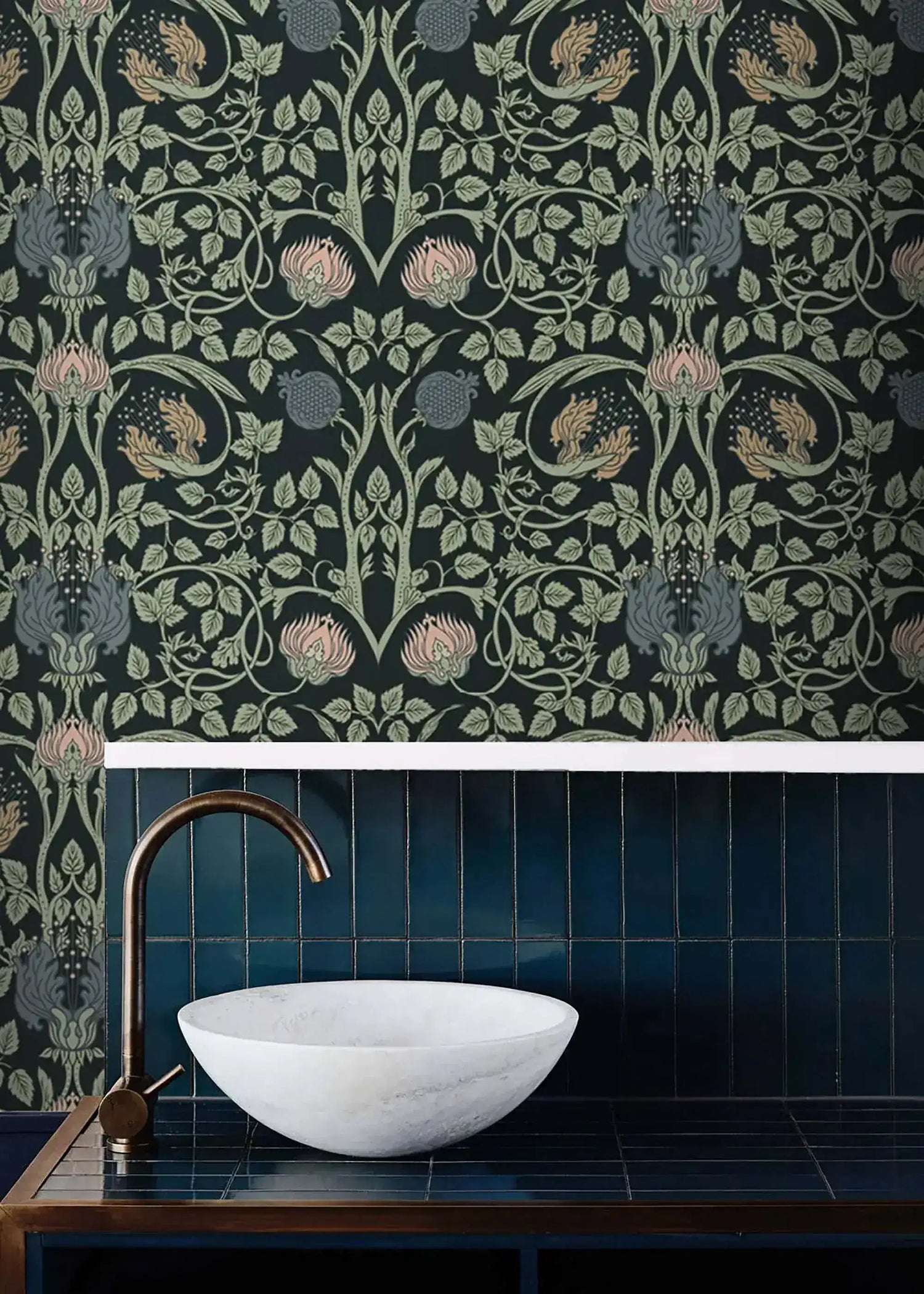 Vine and flower wallpaper in bathroom above dark blue vertical tiles with white marble basin and bronze gooseneck faucet