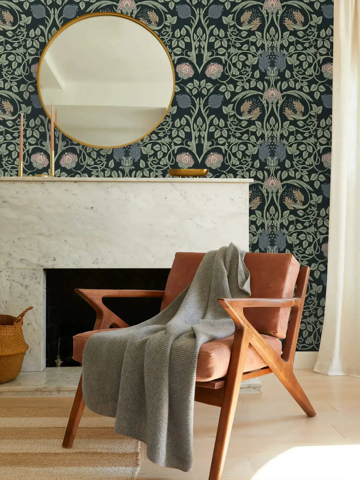 Vine and flower wallpaper around marble fireplace with gold round mirror; mid-century armchair with grey knit throw