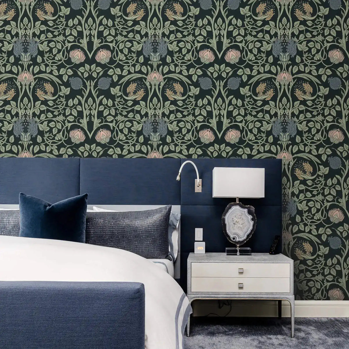 Vine and flower wallpaper behind navy upholstered bed with silver nightstand, agate table lamp, and reading sconce