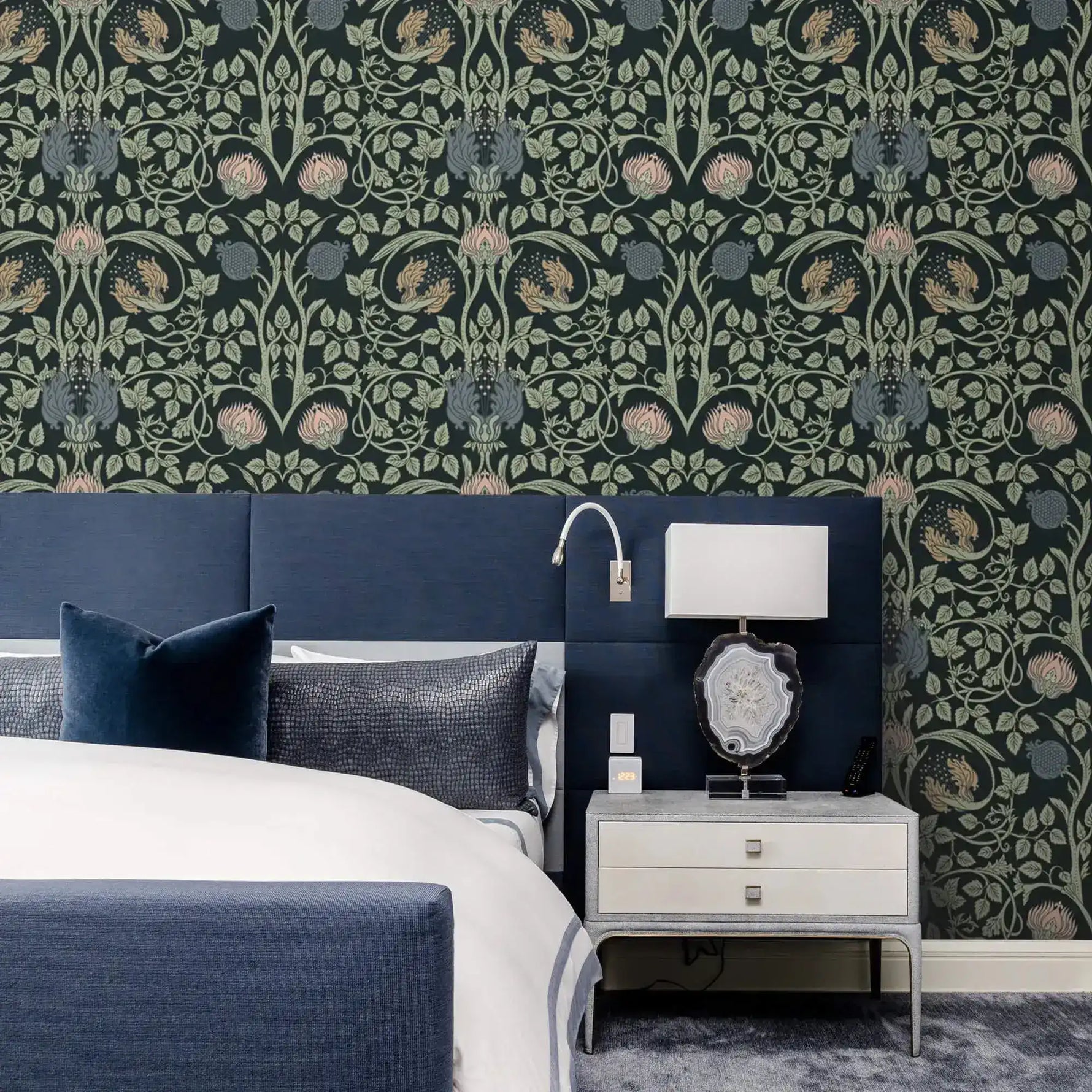 Vine and flower wallpaper behind navy upholstered bed with silver nightstand, agate table lamp, and reading sconce