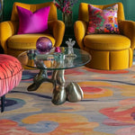 A vibrant and colorful area rug with swirling patterns in a living room setting. The wool rug features a mix of warm and cool tones, adding a lively touch to the space. It is a large, square area rug ideal for bedrooms or living rooms, fitting perfectly under a modern glass coffee table and flanked by mustard yellow armchairs with colorful cushions.