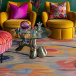 A vibrant and colorful area rug with swirling patterns in a living room setting. The wool rug features a mix of warm and cool tones, adding a lively touch to the space. It is a large, square area rug ideal for bedrooms or living rooms, fitting perfectly under a modern glass coffee table and flanked by mustard yellow armchairs with colorful cushions.