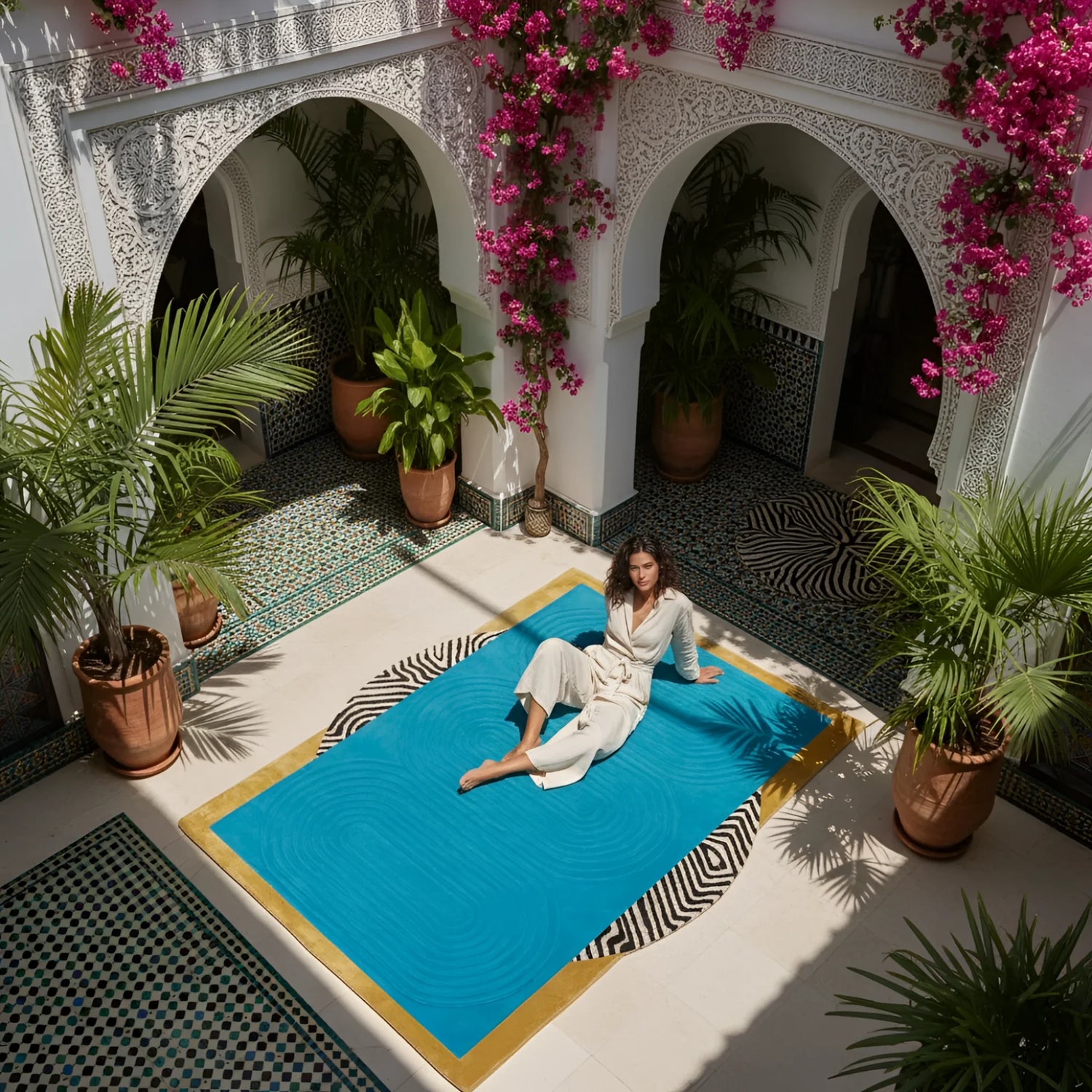 Woman sitting on a blue rug in a courtyard with an abstract wool rug, white walls, arches, and potted plants.