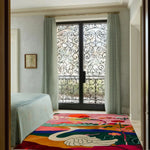 A vibrant area rug with a colorful abstract design featuring a swan, set in a spacious living room. This wool rug adds a touch of elegance, perfect for large living room spaces or an office setting. The room has a beautiful wrought iron window and light-colored curtains complementing the intricate design of the rug.