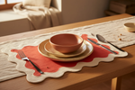 White and red linen scallop placemat set under stacked bowls and gold cutlery on a wooden table.