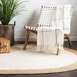 A cozy living room scene featuring a white oval jute area rug. The rug adds a natural touch to the dark hardwood floor, complementing the wicker chair with a white throw. Perfect for those looking for stylish and eco-friendly rugs for their living room.