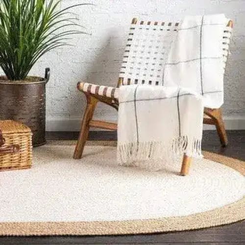 A cozy living room scene featuring a white oval jute area rug. The rug adds a natural touch to the dark hardwood floor, complementing the wicker chair with a white throw. Perfect for those looking for stylish and eco-friendly rugs for their living room.