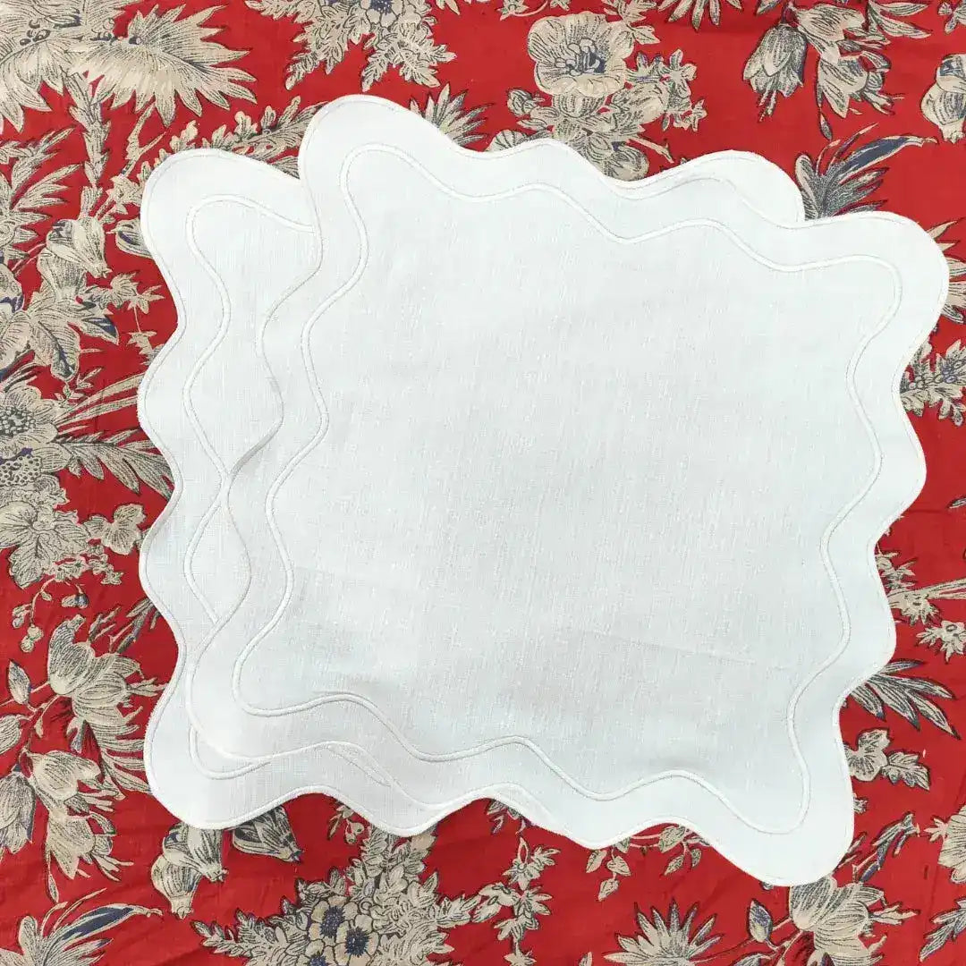 Two white scallop linen square placemats with wavy stitched edge layered on a red floral tablecloth, top view