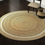 A beautifully crafted round area rug made from natural jute, featuring a white trim that adds a touch of elegance. This boho rug is perfect for a dining room setting, complementing the dark wooden floor and nearby chair. Ideal for formal dining rooms, this jute rug combines rustic charm with modern style.