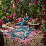 Woman in a colorful dress sitting on a pink and blue patterned rug in a lush garden setting.