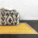 A vibrant yellow rectangular jute area rug placed on a dark wooden floor, perfect for adding a boho touch to bedrooms. This stylish rug, ideal for those seeking rugs for bedroom spaces, is paired with a black and white patterned pouf, complementing the bohemian aesthetic.