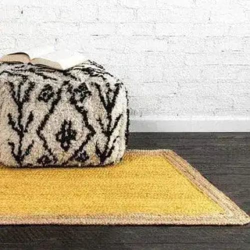 A vibrant yellow rectangular jute area rug placed on a dark wooden floor, perfect for adding a boho touch to bedrooms. This stylish rug, ideal for those seeking rugs for bedroom spaces, is paired with a black and white patterned pouf, complementing the bohemian aesthetic.