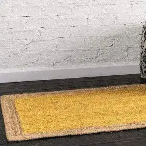 A vibrant yellow rectangular jute area rug placed on a dark wooden floor against a white brick wall. This boho rug is perfect for adding a pop of color to any bedroom, making it an ideal choice for those looking to enhance their space with stylish bedroom area rugs.