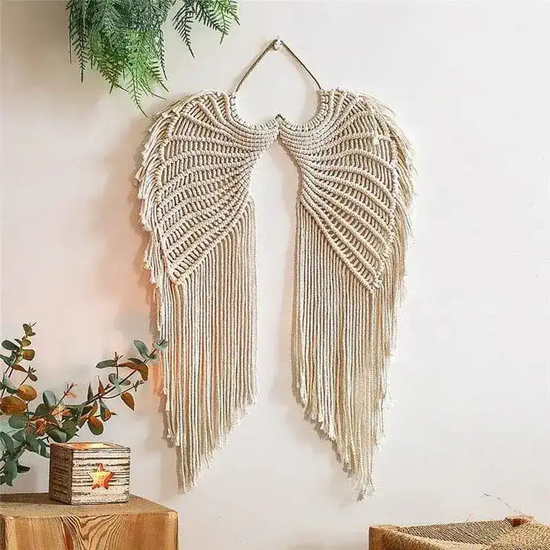 Cream macrame angel wings wall hanging with fringed tassels on a white wall with eucalyptus and fern