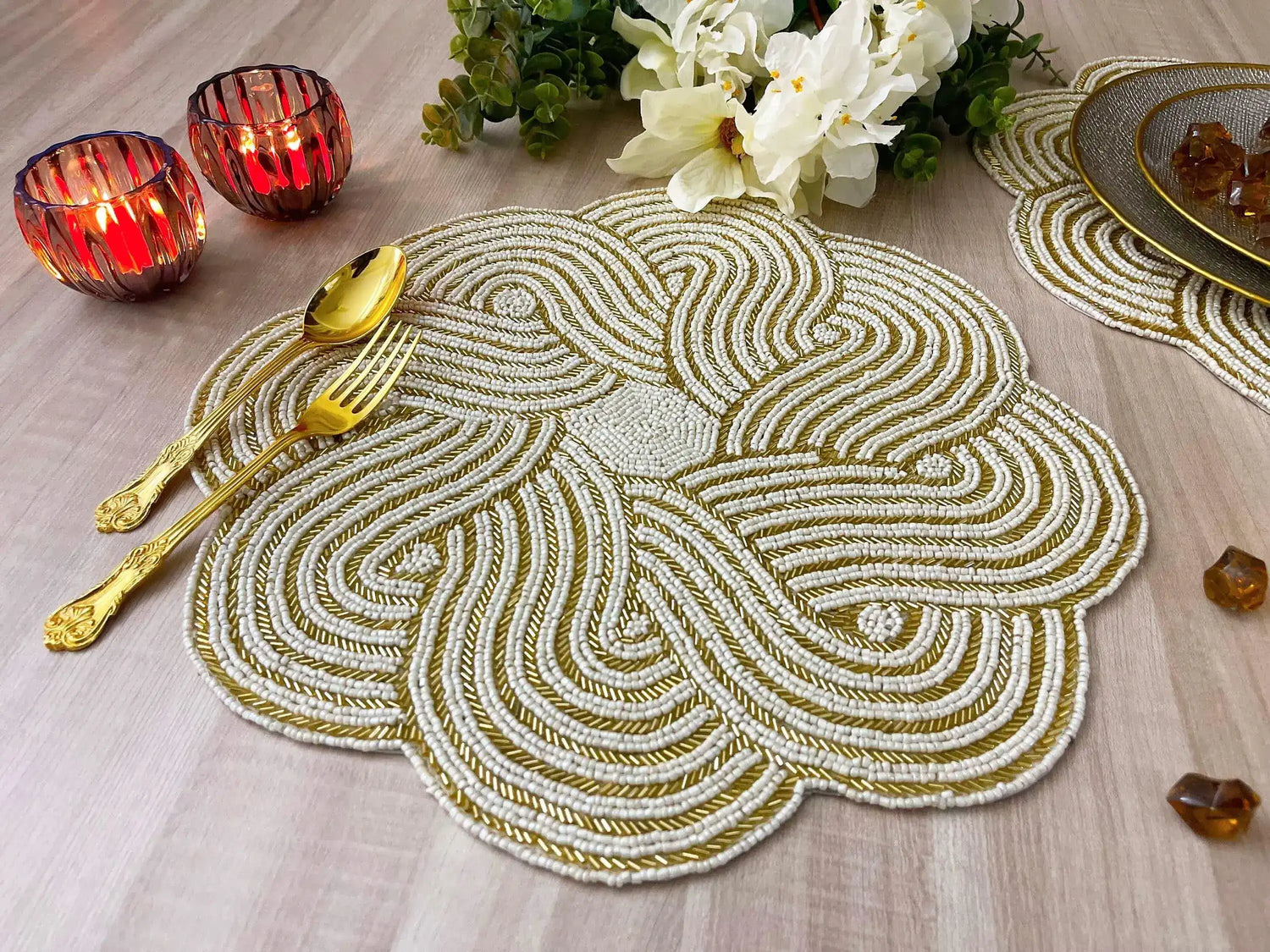 Front view of intertwined scallop placemat in candlelit setting, two red candle holders, white flowers, amber gems