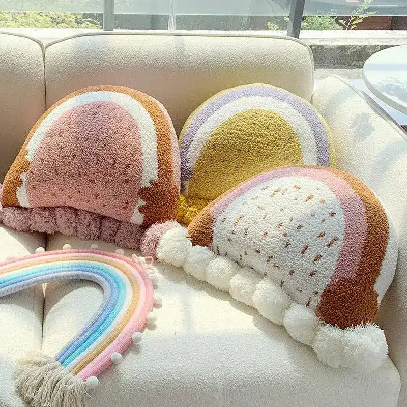 Rainbow-shaped embroidered decorative throw pillows in pastel colors, featuring fluffy pom-pom accents, arranged on a light-colored sofa.