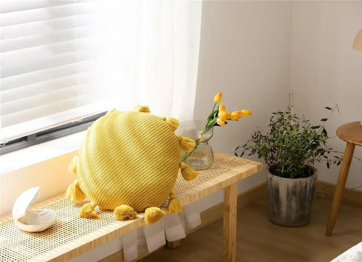 Round tassel decorative throw pillow in vibrant yellow color, placed on a wooden bench with natural light filtering through the window.