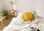 Round tassel decorative throw pillow in sunny yellow color placed on a cozy bed with white bedding, accompanied by a wooden side table and a potted plant.