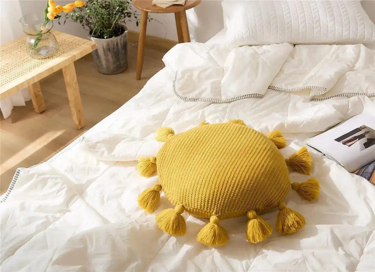 Round yellow decorative throw pillow with tassels on a bed, adding a pop of color and texture to home decor.
