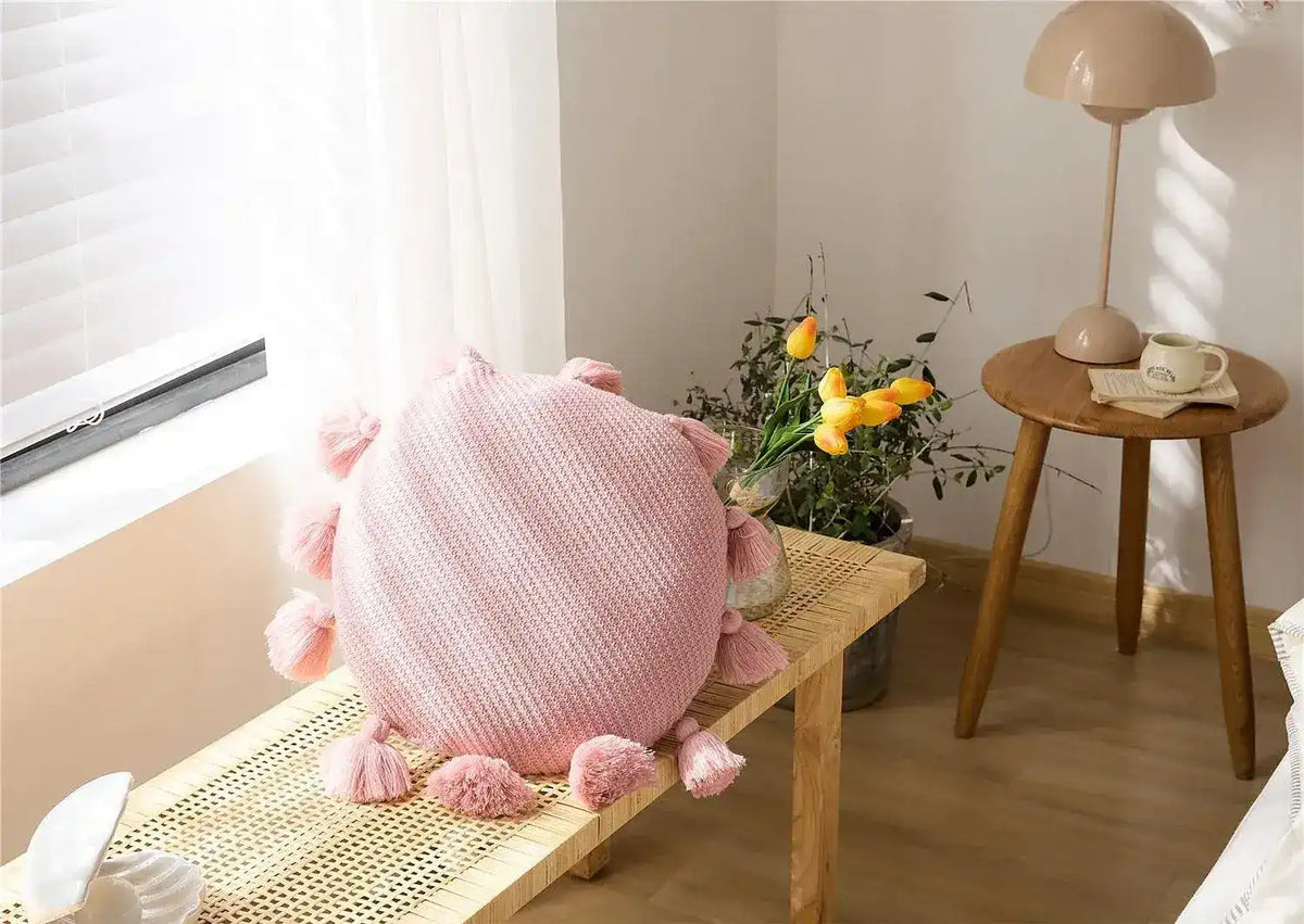 Round pink decorative throw pillow with tassels on a wooden table in a cozy interior setting.