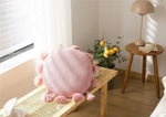 Round pink decorative throw pillow with tassels on a wooden table in a cozy interior setting.