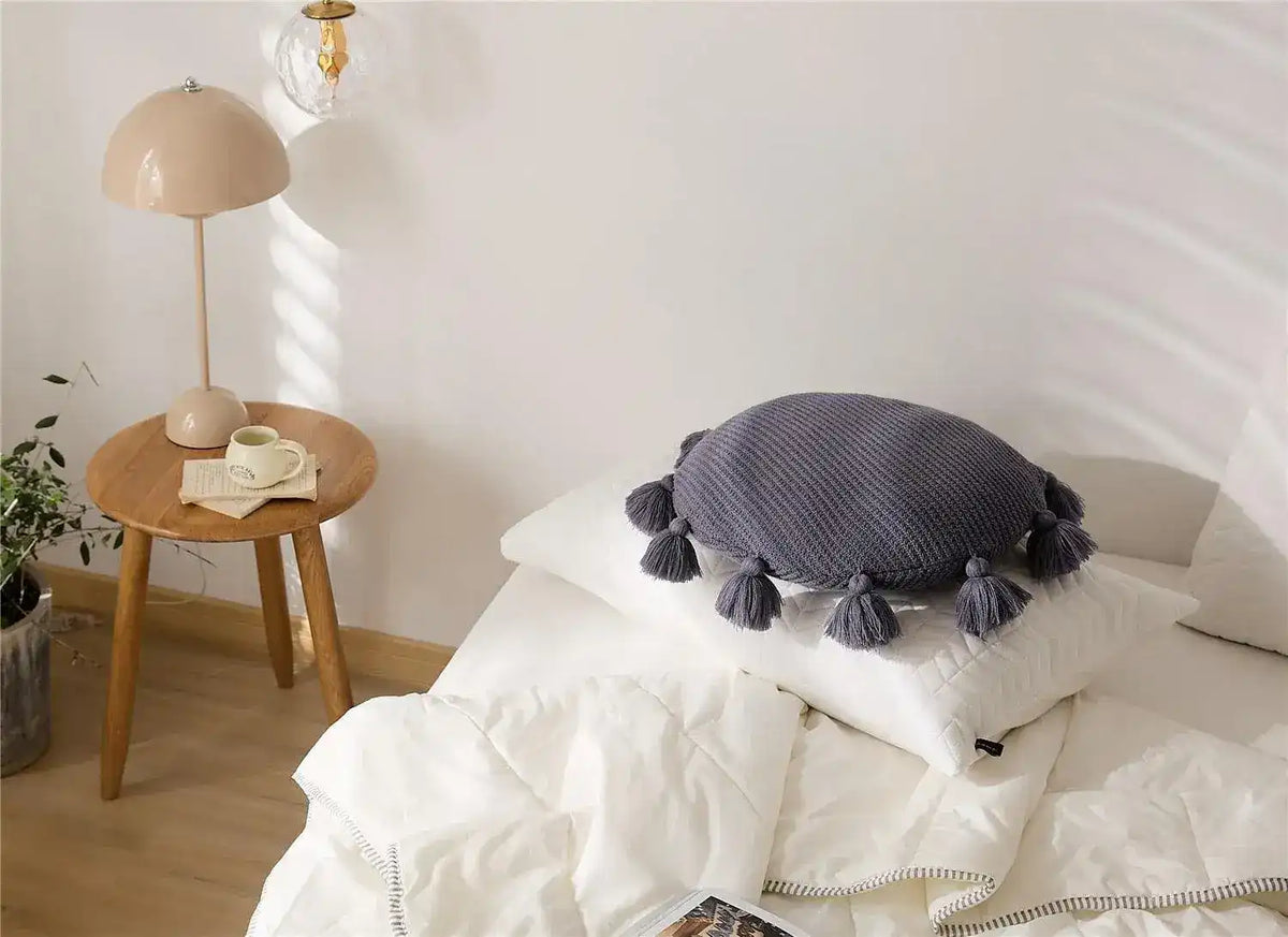 Round decorative throw pillow with tassels placed on a bed, enhancing the cozy interior decor.
