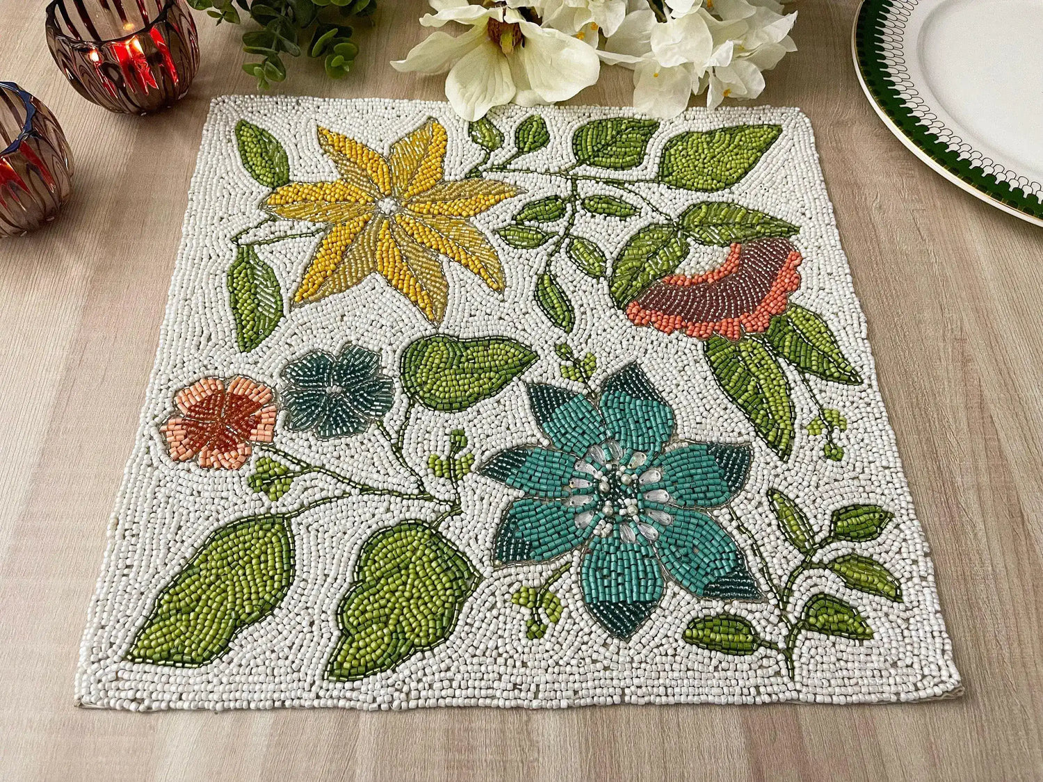 Front view of spring flower square beaded placemat on dining table beside white plate and candles, full design visible